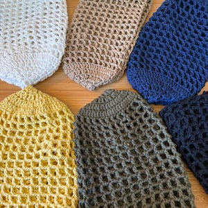 Könnte beinhalten: Sechs gehäkelte Einkaufstaschen in verschiedenen Farben: Weiß, Beige, Marineblau, Gelb, Grau und Dunkelblau. Die Taschen sind oval geformt und haben ein Netzdesign, geeignet für Obst und Gemüse.