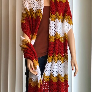 May include: A crocheted scarf in red, white, and gold with a wavy pattern. The scarf is draped over a mannequin.