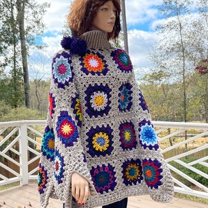 Puede incluir: Un poncho de ganchillo beige con cuello alto y un patrón de cuadrados de la abuela multicolores en tonos azul, amarillo, rojo y morado. La prenda está decorada con dos pompones de lana de color morado oscuro en el escote.