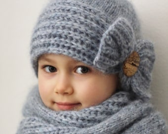May include: A blue knitted hat and scarf set with a large bow on the hat. The hat has a folded brim and a button detail. The scarf is a cowl style. The text "PDF PATTERN" is visible at the bottom of the image.