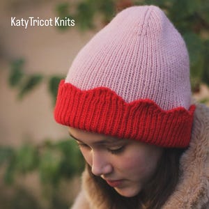 May include: A knitted beanie with a light pink crown and a red scalloped brim. The text "Katy Tricot Knits" is at the top left. The text "PDF PATTERN" and "Holding the Wave Beanie" are at the bottom. The beanie is worn by a person.