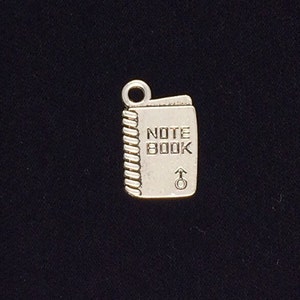 May include: Silver charm shaped like a notebook with the words "NOTE BOOK" on the front. The charm has a loop at the top for attaching to a bracelet or necklace.