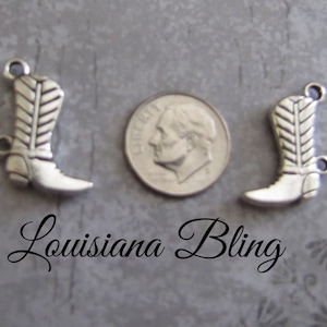 May include: Two silver cowboy boot charms, each with a detailed design, are shown next to a US dime for size comparison. The charms have a loop at the top for attaching to jewelry or other crafts.