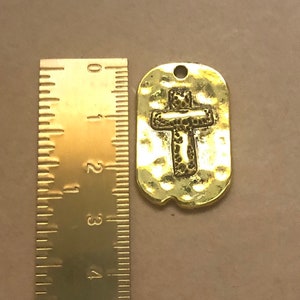 May include: A gold-toned metal charm in the shape of a dog tag with a raised cross design. The cross is textured and has a hammered finish. The charm is approximately 1.5 inches long.