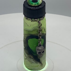 Peut inclure: Petite bouteille décorative avec un bouchon noir et un ornement en forme d'œil vert. La bouteille présente un motif tourbillonnant vert et noir, une chaîne argentée et un charme de crâne. La bouteille mesure environ 7.6 cm de haut.