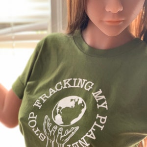 以下が含まれることがあります： 地球儀を持った手を白で描いた緑色のTシャツ。"Stop Fracking My Planet"という文字が入っています。