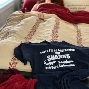 Peut inclure: Un t-shirt bleu marine avec le texte blanc "Sorry I'm so Aggressive but SHARKS Are Born Swimming". Le t-shirt est posé sur un lit avec une couette à motifs et un oreiller rouge. Des vêtements rouges sont visibles au premier plan.