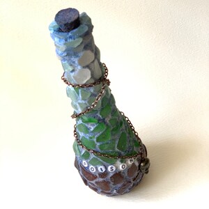 Peut inclure: Une bouteille décorative avec un motif en mosaïque dans les tons de vert, de bleu et de marron. Le mot "POISON" est épelé en perles blanches autour de la base. Une chaîne entoure la bouteille et un bouchon sombre scelle le dessus.
