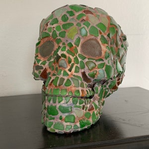 Peut inclure: Une sculpture de crâne décorative faite de morceaux de verre de mer verts et bruns. Le design en mosaïque recouvre tout le crâne, avec des orbites et une mâchoire détaillées. La sculpture est posée sur une surface sombre.