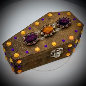 Peut inclure: Une boîte en bois en forme de cercueil avec un couvercle à charnière. La boîte est teintée en marron et décorée de strass orange et violets. Trois grands bijoux ornés dans les tons orange et violet ornent le dessus.