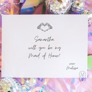 May include: A white card with the text "Samantha, will you be my Maid of Honor?" is surrounded by disco balls and iridescent confetti. The card has a heart-shaped design and is signed "Melissa".