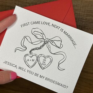 May include: A white card with a black line drawing of a bow and two heart-shaped lockets. The card is inside a red envelope. Text on the card reads: "FIRST CAME LOVE, NEXT IS MARRIAGE... JESSICA, WILL YOU BE MY BRIDESMAID?"