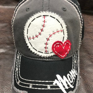 May include: A gray and black baseball cap with a white baseball and red heart applique. The cap has a distressed look and the word "Mom" is embroidered on the brim in white.