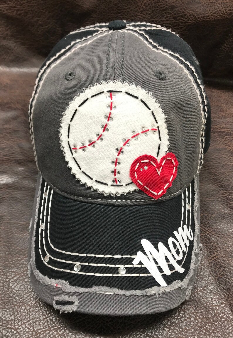 May include: A gray and black baseball cap with a white baseball and red heart applique. The cap has a distressed look and the word "Mom" is embroidered on the brim in white.
