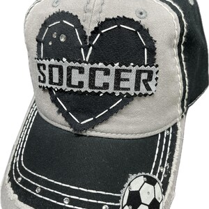 May include: A grey and black baseball cap with a black heart and the word "SOCCER" in silver glitter. A white football with black stitching is on the side of the cap.