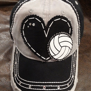 May include: A grey and black baseball cap with a black heart and a white volleyball on the front. The cap has a distressed look and rhinestones around the brim.