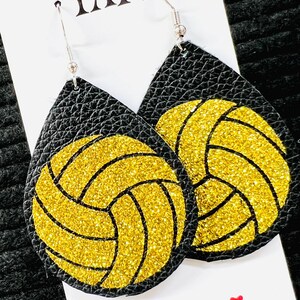May include: Black teardrop earrings with a gold glitter volleyball design. The earrings have silver hooks and are displayed on a white card with the text "YAY!" and "It's Gameday".