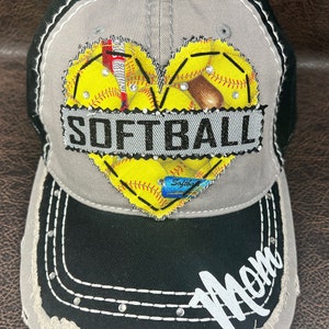 Può includere: Un cappello da baseball grigio e nero con un aspetto usurato. La parte anteriore del cappello presenta un design a forma di cuore con tema softball. Il cuore è fatto di tessuto giallo con cuciture rosse e strass. La parola "SOFTBALL" è scritta in lettere nere su una fascia nera che attraversa il cuore. Il visore del cappello è nero con cuciture bianche e strass. La parola "Mamma" è scritta in lettere bianche sul visore.