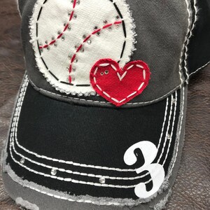 May include: Gray baseball cap with a white baseball and red heart applique. The cap has a black brim and the number 3 is embroidered on the front.