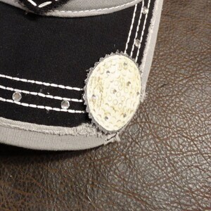 May include: A black and gray baseball cap with a white stitched brim and a white golf ball design on the front.