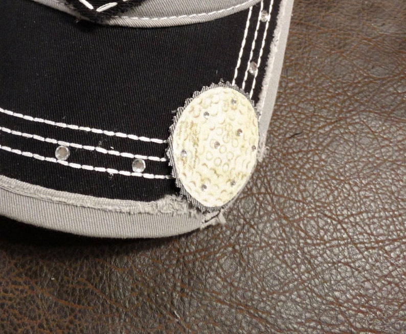 May include: A black and gray baseball cap with a white stitched brim and a white golf ball design on the front.