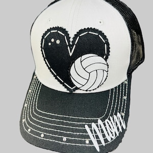 May include: White and black trucker hat with a black heart and a white volleyball. The brim of the hat is black with white stitching and the word "Mom" in white with rhinestones.