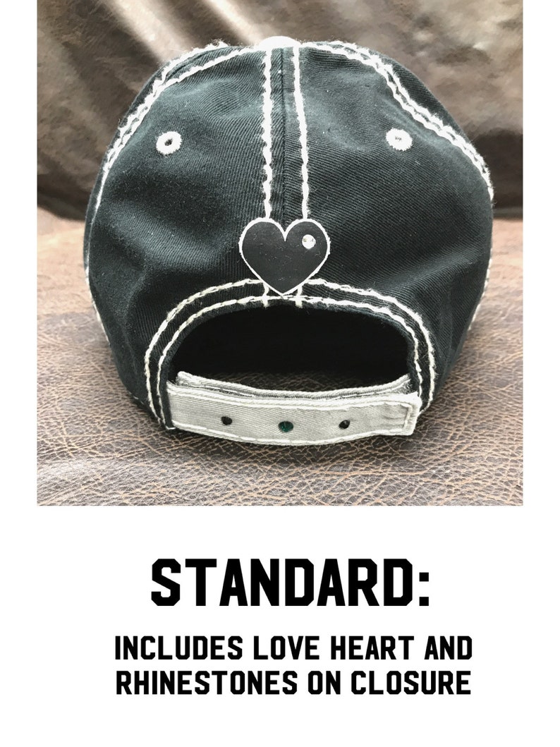 May include: Black baseball cap with white stitching and a black heart with a rhinestone on the back. The cap has a gray adjustable strap with rhinestones.