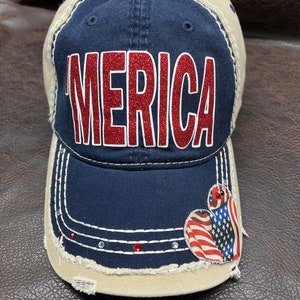 May include: A blue and beige baseball cap with a distressed look. The cap has a red glitter "MERICA" on the front and a red, white, and blue heart-shaped American flag patch on the side.