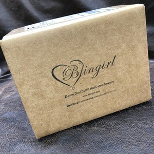 以下が含まれることがあります： 「Blingirl Game Day Spirit Hats and Jewelry」とウェブサイトアドレス「www.Blingirl.com」が印刷された茶色の段ボール箱です。