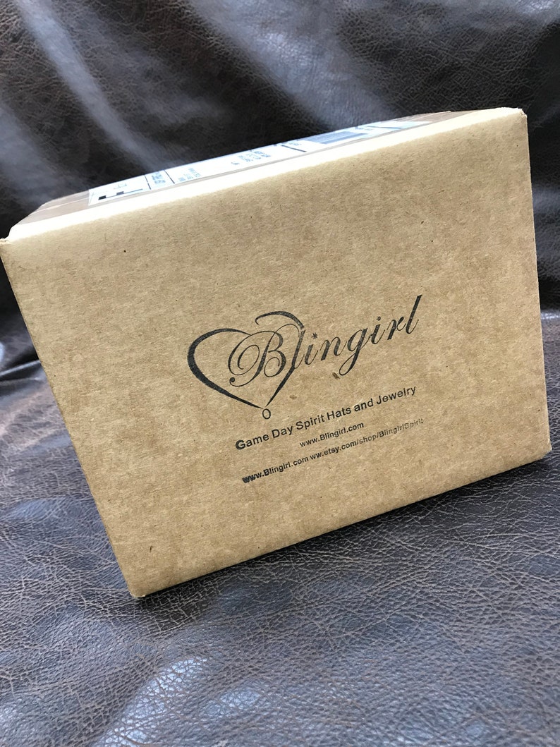 以下が含まれることがあります： 「Blingirl Game Day Spirit Hats and Jewelry」とウェブサイトアドレス「www.Blingirl.com」が印刷された茶色の段ボール箱です。