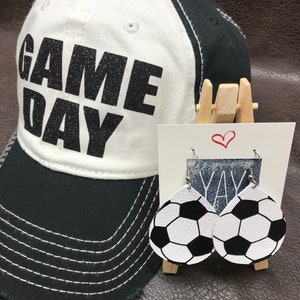 Peut inclure: Une casquette de baseball blanche et noire avec le texte "GAME DAY" en paillettes noires. Une paire de boucles d'oreilles en cuir blanc avec un motif de ballon de football noir.