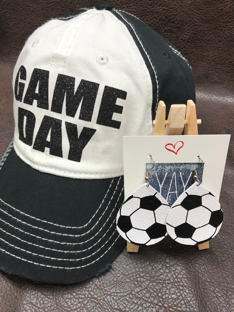 Peut inclure: Une casquette de baseball blanche et noire avec le texte "GAME DAY" en paillettes noires. Une paire de boucles d'oreilles en cuir blanc avec un motif de ballon de football noir.