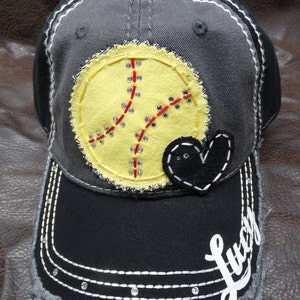 May include: A black and gray baseball cap with a yellow softball design, a black heart, and the word "Lucy" in white lettering.