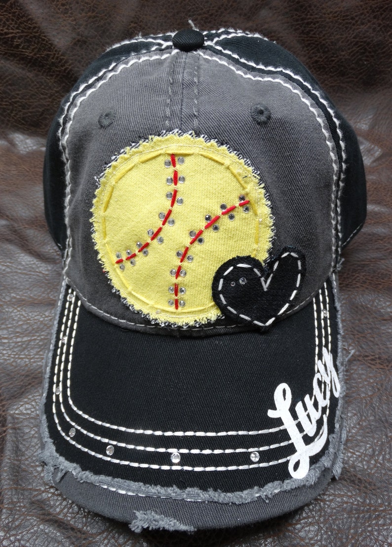 May include: A black and gray baseball cap with a yellow softball design, a black heart, and the word "Lucy" in white lettering.