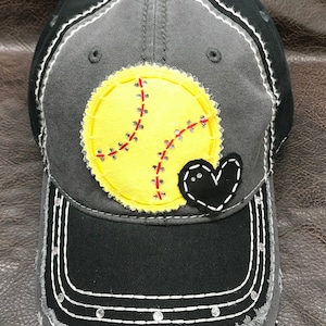 May include: A gray and black baseball cap with a yellow softball and a black heart sewn on the front. The cap has a distressed look with white stitching and rhinestones around the brim.