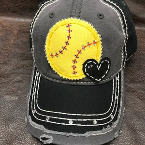 May include: A gray and black baseball cap with a yellow softball and a black heart sewn on the front. The cap has a distressed look with frayed edges and rhinestones on the brim.