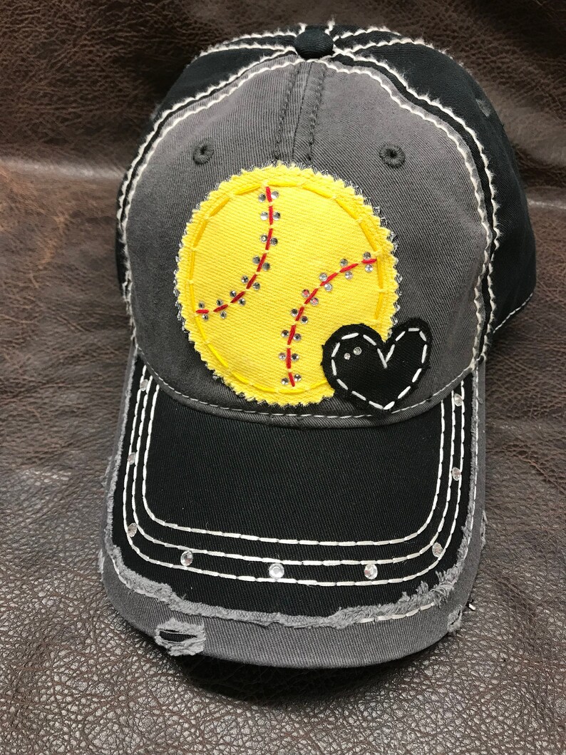 May include: A gray and black baseball cap with a yellow softball and a black heart sewn on the front. The cap has a distressed look with frayed edges and rhinestones on the brim.