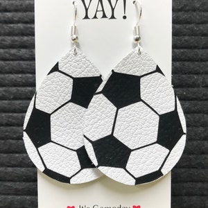 Peut inclure: Une paire de boucles d'oreilles en cuir blanc en forme de ballon de football avec des coutures noires. Les boucles d'oreilles sont suspendues &agrave; des crochets argent&eacute;s et sont pr&eacute;sent&eacute;es sur une carte blanche avec le texte "YAY!" et "It's Gameday".