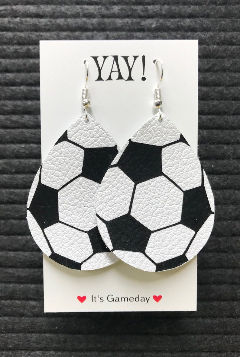 Peut inclure: Une paire de boucles d'oreilles en cuir blanc en forme de ballon de football avec des coutures noires. Les boucles d'oreilles sont suspendues &agrave; des crochets argent&eacute;s et sont pr&eacute;sent&eacute;es sur une carte blanche avec le texte "YAY!" et "It's Gameday".