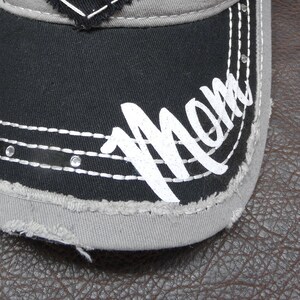 May include: A black and gray baseball cap with a distressed look. The cap has white stitching and the word "Mom" written in white on the front.