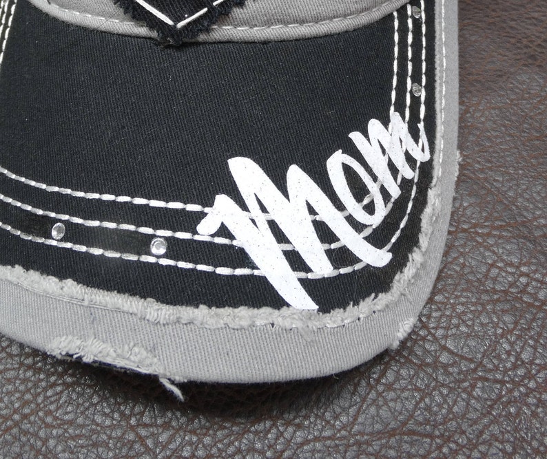 May include: A black and gray baseball cap with a distressed look. The cap has white stitching and the word "Mom" written in white on the front.