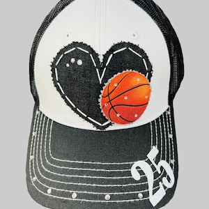 May include: A black and white trucker hat with a basketball theme. The front panel features a black heart with a basketball and the number 25. The hat is embellished with rhinestones.