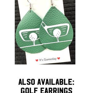 May include: A pair of green leather teardrop-shaped earrings with a white golf club and ball design. The earrings have silver hooks. The text "Yay!" and "It's Gameday" are visible on the white card behind the earrings.