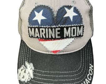 May include: A gray and black trucker hat with a blue and white heart design that says "MARINE MOM" on it. The hat has a black bill with a silver Marine Corps emblem on the left side and the word "CHACON" embroidered on the right side.