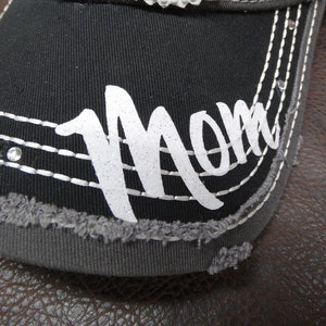 May include: Black baseball cap with a gray brim and white glitter lettering that says "Mom".