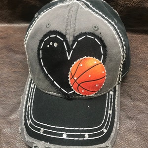 May include: A gray and black baseball cap with a black heart and an orange basketball on it. The heart has rhinestones on it.