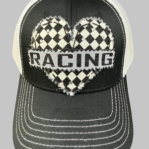 May include: A black and white trucker hat featuring a frayed-edge heart patch with a black and white checkered pattern. The word RACING is printed in black glittery letters across a light gray horizontal bar overlaying the center of the heart.