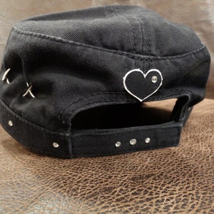 May include: Black cotton baseball cap with a black heart patch and rhinestones. The heart patch has a white outline and a single rhinestone in the centre.