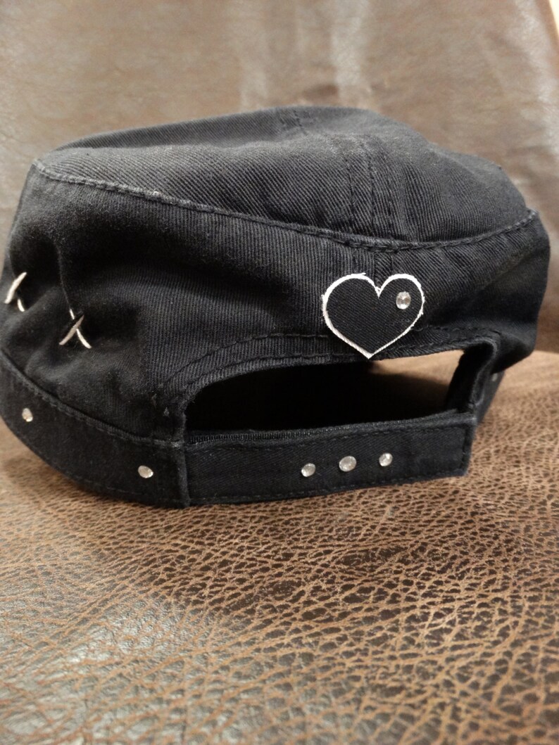 May include: Black cotton baseball cap with a black heart patch and rhinestones. The heart patch has a white outline and a single rhinestone in the centre.