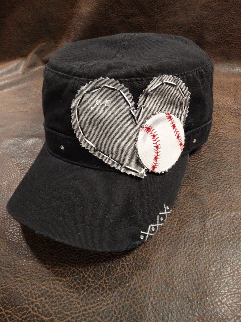 May include: A black baseball cap with a white heart-shaped patch and a baseball sewn on. The heart has stitching around the edges and rhinestones in the centre.